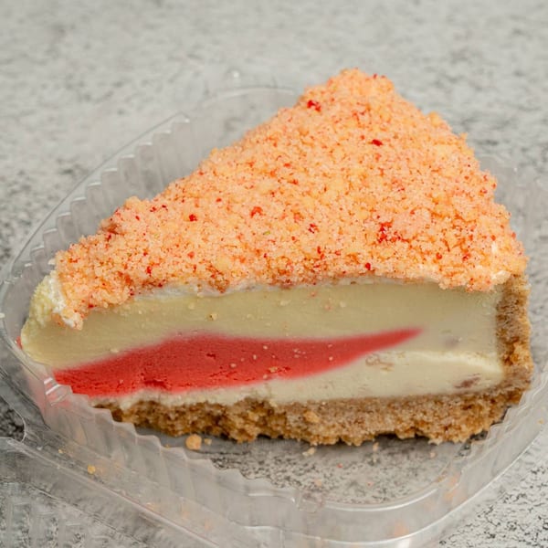 A slice of cheesecake with a graham cracker crust, red swirl, and orange crumble topping in a plastic container.