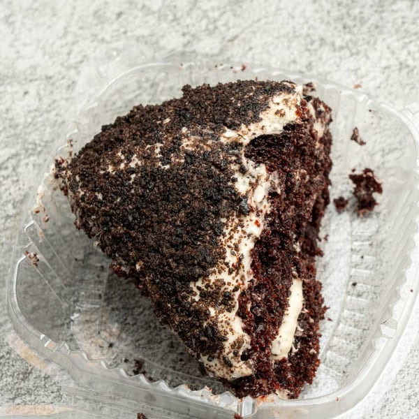 A slice of Angie's Soul House chocolate cake with white frosting and chocolate crumbles in a plastic container.